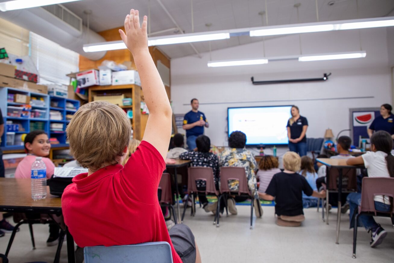 A boy in a red shirt raises his hand during a career day presentation