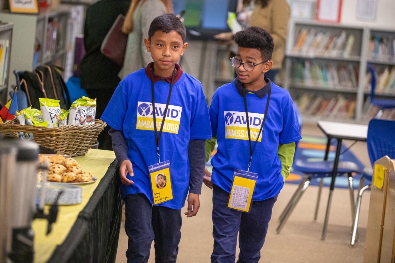 Two boys in blue AVID Revealed shirts and lanyards stand next to the refreshments table