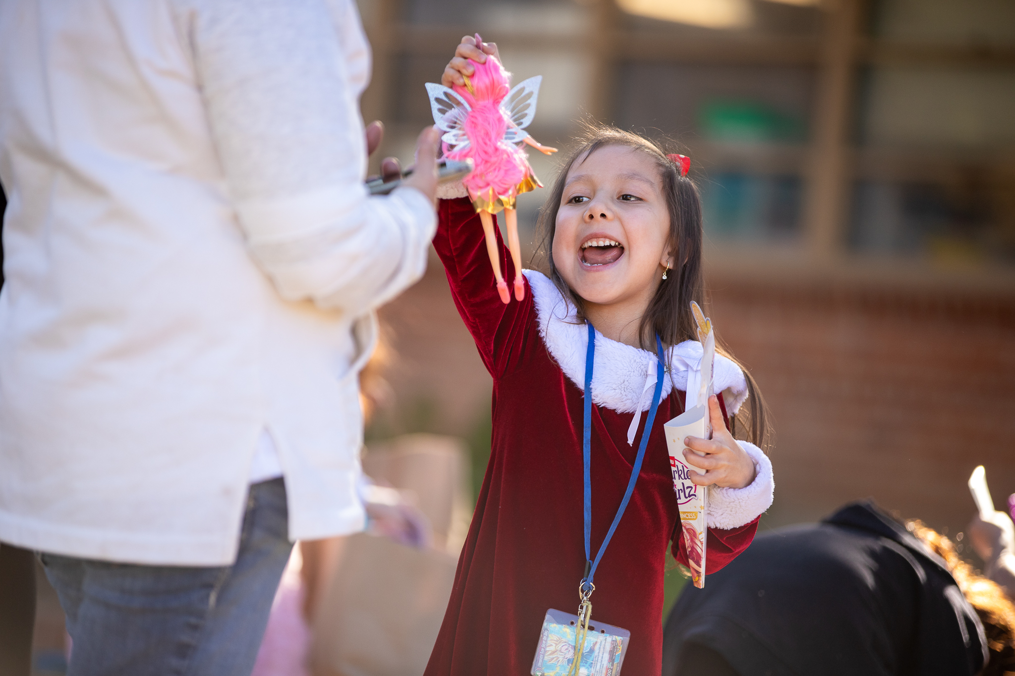 A little girl in a Santa dress smiles as she holds up her new fairy doll