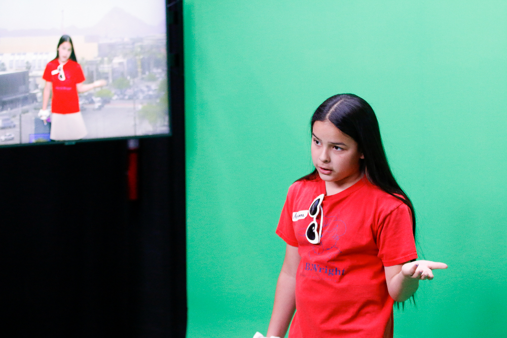 A girl in a red shirt acts out a weather cast in front of the green screen
