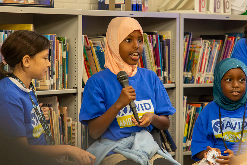 A girl in a blue AVID Ambassador shirt and peach headscarf talks into a microphone