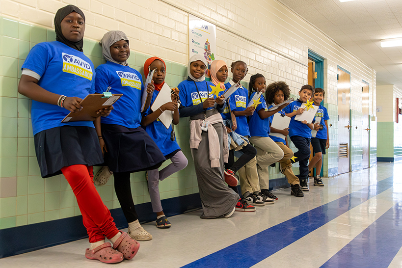 A group of students in blue AVID Ambassador shirts stand in the hallway holding clipboards