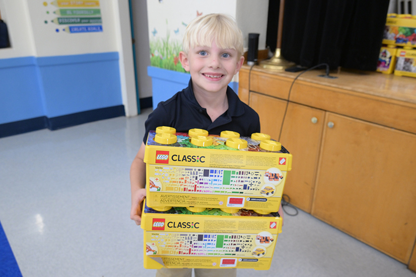 A little boy with light blonde hair smiles while holding up his Lego kit
