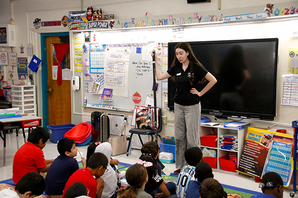 A woman holds a hockey stick as she talks to a classroom of students